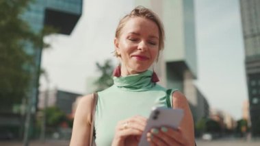 Beautiful woman with short blond hair in casual clothes is using cellphone on modern building background
