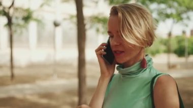 Close up, beautiful woman with short blond hair in casual clothes walks through the city square, talking on cellphone