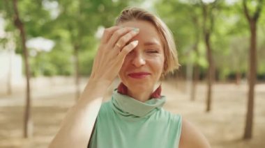 Close-up of happy woman with short blond hair in casual clothes, fixing her hair, smiling at the camer