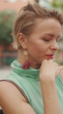 VERTICAL VIDEO: Beautiful woman with short blond hair in casual clothes sits on bench, smiles, enjoys warm summer day