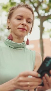 VERTICAL VIDEO: Beautiful smiling woman with short blond hair in casual clothes sits on bench, uses cellphone