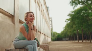 Beautiful woman with short blond hair in casual clothes sits on bench, smiles, enjoys warm summer day