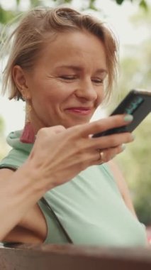 VERTICAL VIDEO: Beautiful smiling woman with short blond hair in casual clothes sits on bench, sends voice message on cellphone