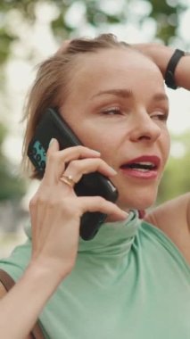 VERTICAL VIDEO: Close up, beautiful happy smiling woman with short blond hair in casual wear sits on bench, talking on cellphone