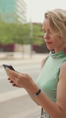 VERTICAL VIDEO: Beautiful woman with short blond hair in casual clothes sits at public transport stop, uses mobile phone
