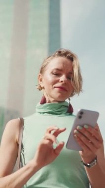 VERTICAL VIDEO: Beautiful woman with short blond hair in casual clothes is using cellphone on modern building background