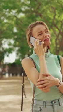 VERTICAL VIDEO: Beautiful smiling woman with short blond hair in casual clothes walks through the city square, talking on cellphone