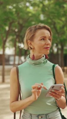 VERTICAL VIDEO: Beautiful woman with short blond hair in casual clothes walks through the city square, uses cellphone