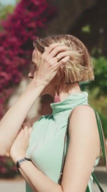VERTICAL VIDEO: Attractive happy woman with short blond hair wearing casual clothes stands at the crosswalk, turns her head and smiles