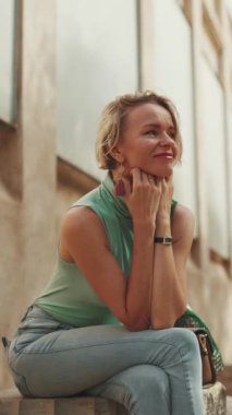 VERTICAL VIDEO: Beautiful woman with short blond hair in casual clothes sits on bench, smiles, enjoys warm summer day