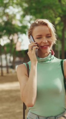 VERTICAL VIDEO: Beautiful smiling woman with short blond hair in casual clothes walks through the city square, talking on cellphone