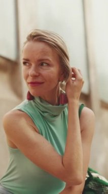 VERTICAL VIDEO: Beautiful woman with short blond hair in casual clothes sits on bench, smiles, enjoys warm summer day