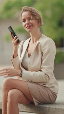 VERTICAL VIDEO: Businesswoman with blond hair talking on cellphone, looking at wrist watch