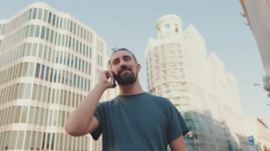 Smiling young man with beard talking on the phone on modern buildings background