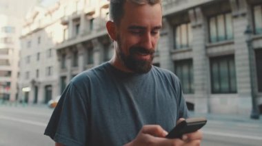 Young smiling man with beard stands on street using cellphone