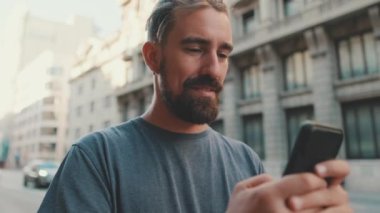 Young smiling man with beard uses smartphone on the road background, turns his head and looks at the camera