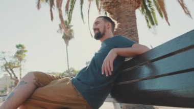 Young smiling man with beard sits and rests on bench. Backlight