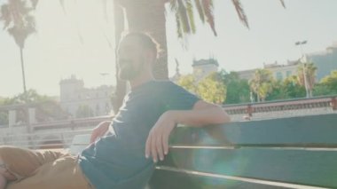 Young smiling man with beard sits and rests on bench. Backlight