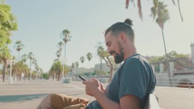 Young smiling man with beard sits on bench , uses cellphone