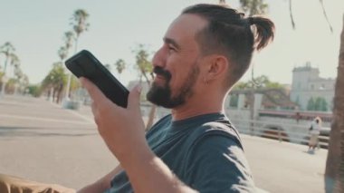 Close-up of happy young man with beard sitting on bench emotionally talking on cellphone on speakerphone