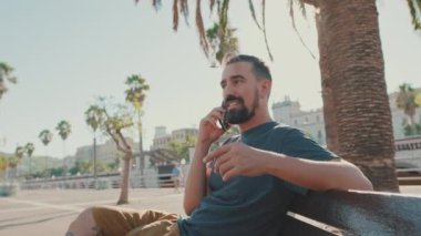 Happy young man with beard sits on bench talking on cellphone