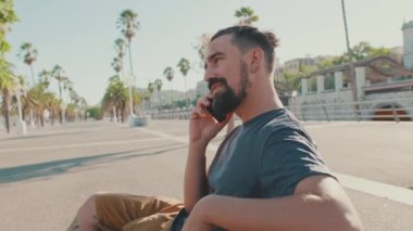 Young smiling man with beard sits on bench, uses cellphone. Backlight