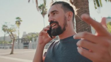 Young man with beard sits on bench emotionally talking on cellphone. Backlight