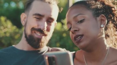 Close up, happy smiling interracial couple watching video on mobile phone while sitting on bench