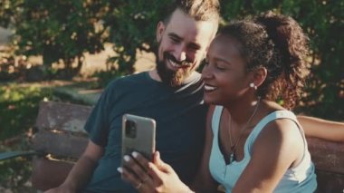 Happy smiling interracial couple making video call on cellphone while sitting on bench