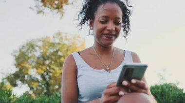 Young mixed race woman sits on park bench, uses cellphone