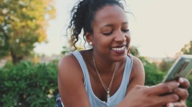 Close up, smiling young mixed race woman sitting on park bench using cellphone