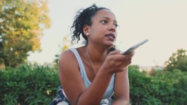 Smiling young mixed race woman sitting on park bench sending voice message