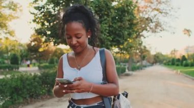 Laughing young mixed race woman walking along path in the park, using cellphone