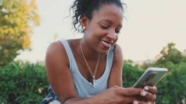 Close up, smiling young mixed race woman sitting on park bench using cellphone. Raises head looking at camera
