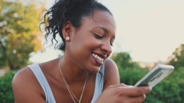 Close-up of laughing young mixed race woman sitting on park bench, sending voice message