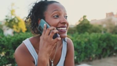 Close-up of laughing young mixed race woman sitting on park bench talking on cellphone