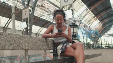 Young mixed race woman sitting at the railway station, using cellphone
