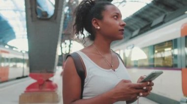 Young mixed race woman stands at the railway station on the background of trains, uses smartphone