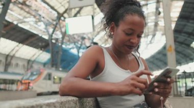 Young mixed race woman sitting at the railway station, using cellphone