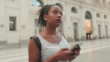 Young mixed race woman stands at the railway station, looks at the information board, uses cellphone