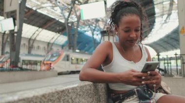 Young mixed race woman sitting at the railway station, using cellphone