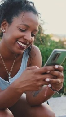 VERTICAL VIDEO: Young mixed race woman sits on park bench, uses cellphone