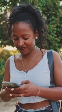 VERTICAL VIDEO: Laughing young mixed race woman walking along path in the park, using cellphone