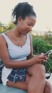 VERTICAL VIDEO: Young mixed race woman sits on park bench, uses cellphone
