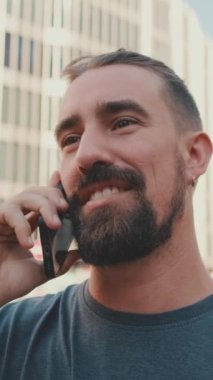 VERTICAL VIDEO: Close-up of young man with beard talking on the phone on traffic cars background