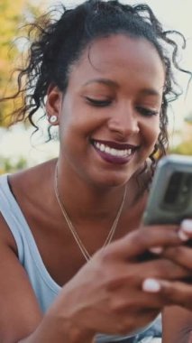 VERTICAL VIDEO: Young mixed race woman sits on park bench, uses cellphone