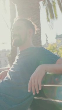VERTICAL VIDEO: Young smiling man with beard sits and rests on bench. Backlight