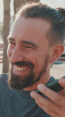 VERTICAL VIDEO: Close-up of young man with beard sitting on bench talking on cellphone on speakerphone