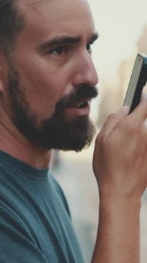 VERTICAL VIDEO: Close-up young man with beard uses cellphone while standing on crosswalk, sends voice message