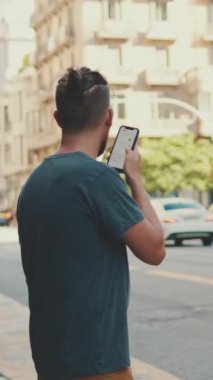 VERTICAL VIDEO: Young man with beard uses cellphone while standing on crosswalk, sends voice message